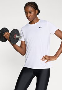 T-shirt sportif blanc à manches courtes avec logo noir, associé à des leggings noirs. Le sujet tient un haltère noir à poignée métallique.