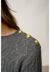 Szary sweter pleciony z teksturowanym wzorem w warkocze, z czterema złotymi dekoracyjnymi guzikami na lewym ramieniu.
