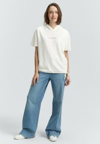 Witte hoodie met een trekkoord en de tekst "goede tijden", gecombineerd met lichtblauwe wijde jeans en witte sneakers tegen een neutrale achtergrond.