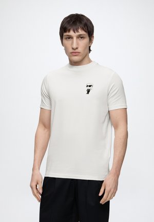 Jeune homme portant un t-shirt blanc avec une petite figure de dessin animé en noir et blanc sur la poitrine, assorti à un pantalon noir, sur un fond uni.