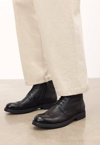 Botine din piele neagră, la glezna, cu detalii brogue, talpă plată și sistem de șireturi, asociate cu pantaloni bej, pe o suprafață netedă.