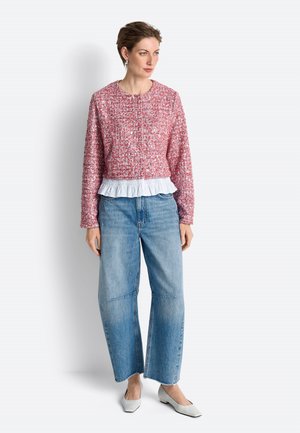 Woman wearing a red textured jacket over a white ruffled blouse, loose blue jeans, and white low-heeled shoes, standing against a white background.