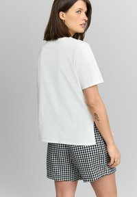 Witte top met korte mouwen en een ontspannen pasvorm, gecombineerd met zwart-wit geruite shorts. De top heeft zijsplitten en een gladde textuur.