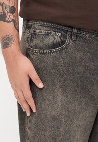 Jeans din denim gri închis cu o textură ușor uzată, având un design tradițional cu cinci buzunare și accente din cupru.