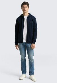 Marineblauer Reißverschluss-Hoodie mit Kapuze, gerippter Textur, frontalen Taschen und goldenen Beschlägen. Kombiniert mit hellblauen Jeans und weißen Turnschuhen.