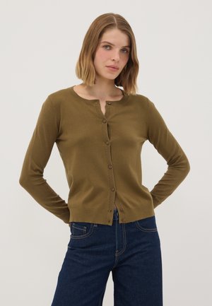 Junge Frau mit schulterlangem blondem Haar, die eine olivgrüne, geknöpfte Strickjacke und dunkelblaue Jeans vor einem einfarbigen Hintergrund trägt.