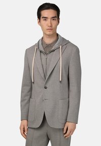 Giacca da abito grigia in tessuto con cappuccio, dotata di due tasche frontali e coulisse. Indossata sopra una camicia grigia abbinata. Finitura testurizzata.