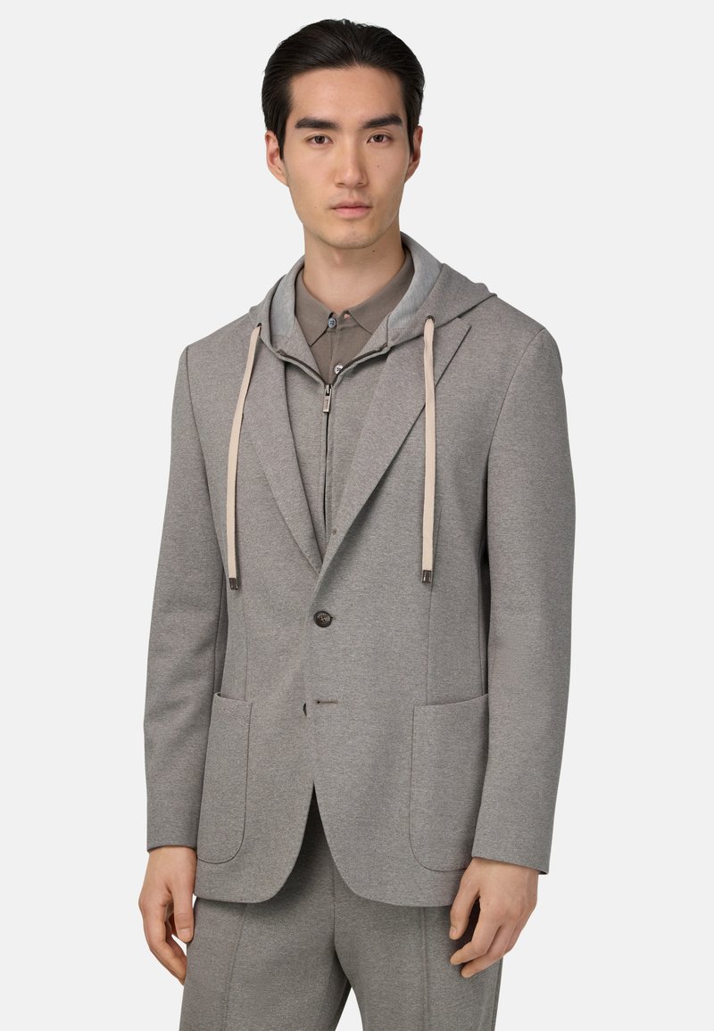 Giacca da abito grigia in tessuto con cappuccio, dotata di due tasche frontali e coulisse. Indossata sopra una camicia grigia abbinata. Finitura testurizzata.