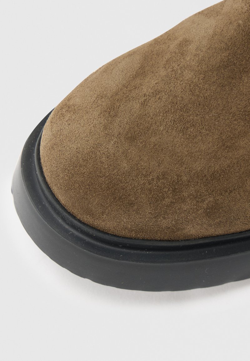 Tomaia di scarpa in camoscio marrone con punta arrotondata e suola in gomma nera. La texture appare morbida e liscia, evidenziando le cuciture lungo i bordi.