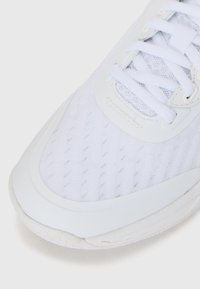 Zapato deportivo blanco con parte superior de malla texturizada, superposiciones sintéticas suaves y cordones planos blancos, con una suela de goma acolchada.