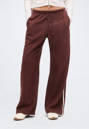 Személy laza, barna, oldalsávos, zsinóros nadrágot és fehér sportcipőt visel, sima világos háttér előtt.