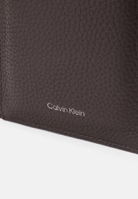 Ciemnobrązowy skórzany portfel o teksturowanej powierzchni i wytłoczonym po boku srebrnym logo "Calvin Klein". Kompaktowy design z starannym przeszyciem.