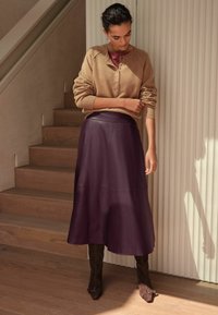 Cárdigan de punto beige con botones, combinado con una falda larga de cuero morado oscuro y botas marrones hasta la rodilla. Escalera de madera de fondo.