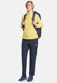 Haut polaire jaune zippé, pantalon cargo bleu marine, chaussures de sport bleues, et un sac à dos noir avec des bretelles. Tenue d'extérieur moderne et fonctionnelle.