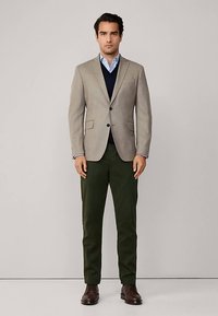 Beige blazer over een navy trui, blauwe blouse, groene broek en bruine schoenen. Het blazer heeft twee voorzakken en twee knopen.