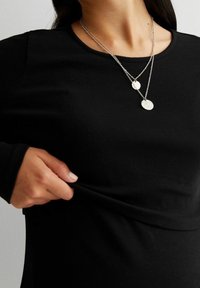 Czarny prążkowany top z długim rękawem o dopasowanym kroju. Dwie warstwowe srebrne naszyjniki z teksturowanymi okrągłymi wisiorkami. Ręka chwytająca materiał.