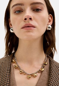 Sølv- og guldhjertevedhængs halskæde på sølvkæde; model iført store sølv kugleøreringe; struktureret brun houndstooth blazer.