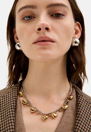 Srebrny i złoty wisiorek w kształcie serca na srebrnym łańcuszku; modelka nosi duże srebrne kolczyki w kształcie kul; strukturalna brązowa marynarka w pepitkę.