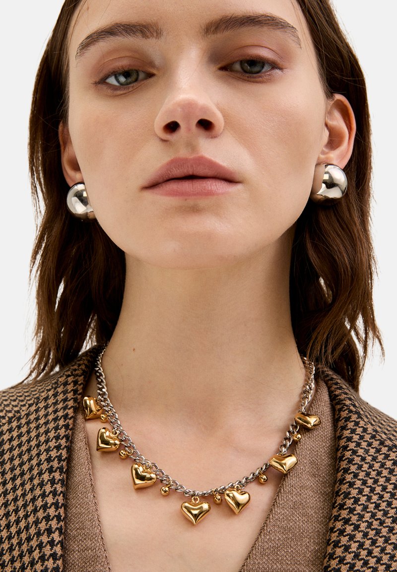 Sølv- og guldhjertevedhængs halskæde på sølvkæde; model iført store sølv kugleøreringe; struktureret brun houndstooth blazer.