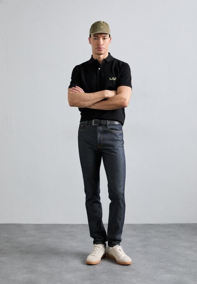 Camisa polo negra con logo dorado, jeans de mezclilla oscuros y zapatos claros. Gorra oliva con logo. El modelo está de pie con los brazos cruzados contra un fondo texturizado.