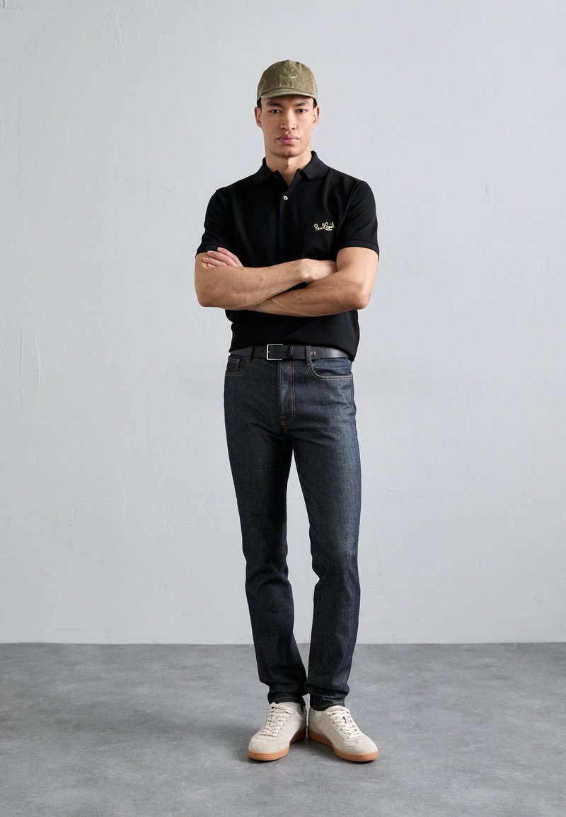 Črna polo majica z zlatim logotipom, temno jeans hlače in svetli čevlji. Olivna kapa z logotipom. Model stoji s prekriženimi rokami ob teksturirani ozadju.