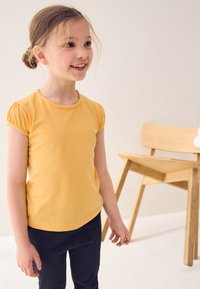 Gelbes Kurzarmoberteil mit gerafften Ärmeln, kombiniert mit schwarzer Hose. Einfacher, glatter Stoff, figurbetontes Design und abgerundeter Ausschnitt.