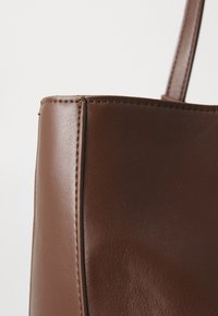 Tote bag in pelle marrone con una texture liscia, evidenti cuciture laterali e una forma strutturata. Presenta un manico abbinato per il trasporto.