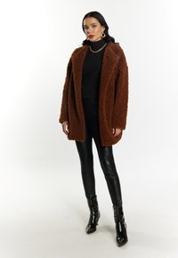 Brun, fuzzy oversized frakke, sort turtleneck, stramme sorte bukser og skinnende sorte ankelstøvler. Accessories inkluderer gyldne hoops øreringe og en kædehalskæde.