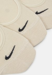 Bež Nike čarape s crnim izvezenim swoosh logotipom. Pleteni materijal i elastičan vrh. Prikazane su tri para, otkrivajući suptilne detalje.
