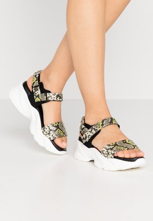 Jambes portant des sandales à semelle blanche épaisse avec des brides à motif peau de serpent noir et jaune sur un sol gris clair.