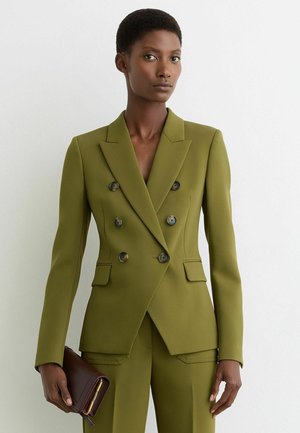 Donna di colore che indossa un blazer doppiopetto verde oliva e pantaloni abbinati, con in mano una pochette marrone su sfondo bianco.