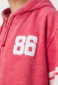Sudadera con capucha roja, con una textura desvanecida y el número "86" bordado en blanco en el pecho. Presenta rayas blancas en las mangas.