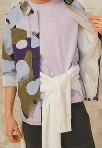 Osoba v purpurovém tričku Converse, vícbarevném sakem s knoflíky a vyhrnutými rukávy, černých kalhotách a bílém svetru svázaném v pase.