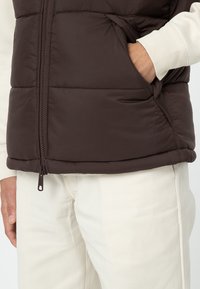 Brun vadderad väst med dragkedja fram och sidoficka. Bärs över en ljusfärgad långärmad topp och beige byxor. Strukturerad tyg.