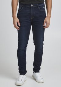 Jeans ajustados de mezclilla azul oscuro con costuras amarillas. Presenta bolsillos delanteros, un bolsillo para monedas y un único bolsillo trasero. Combinados con zapatillas blancas.