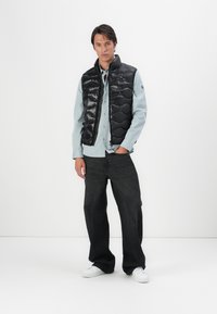 Gilet imbottito nero con finitura lucida, design a ondine, indossato sopra una camicia azzurro chiaro con bottoni, abbinato a jeans scuri e sneakers bianche.