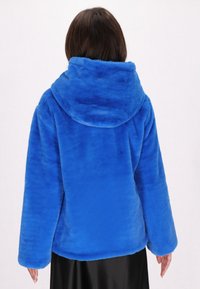 Felpa blu in pelliccia sintetica con un ampio cappuccio e maniche larghe. Tessuto morbido e vestibilità rilassata, che arriva fino a metà fianchi. Orlo della gonna nera visibile sotto.