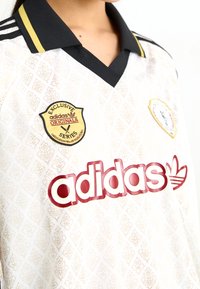 Bílý dres Adidas se černým límcem a žlutými akcenty, složitý listový vzor a logo nášivky černé a červené barvy na hrudi.