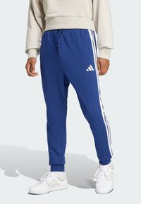 Pantalones de chándal azules hechos de un tejido suave con rayas laterales blancas. Cuentan con una cinturilla elástica y puños en los tobillos, con un detalle de logo.