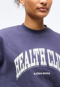 Κοντινό πλάνο ενός ατόμου που φοράει μωβ φούτερ με λευκό κείμενο «HEALTH CLUB» και «BJÖRN BORG» κάτω από το κυρτό σχέδιο.