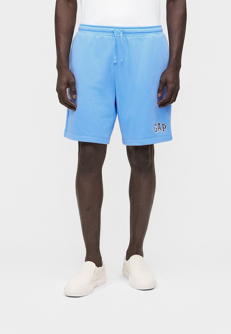 Άνδρας που φοράει ανοιχτό μπλε σόρτς με κορδόνι και λογότυπο "GAP" στο αριστερό πόδι και λευκά παπούτσια slip-on, στέκεται μπροστά σε λευκό φόντο.