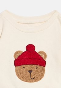 Beiges Babyshirt mit einem bestickten Bärengesicht, das eine rote gestrickte Mütze mit Bommel trägt.
