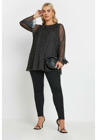 Blusa nera a pois con materiale trasparente e maniche a campana, abbinata a jeans skinny neri e una borsa a tracolla nera intrecciata.