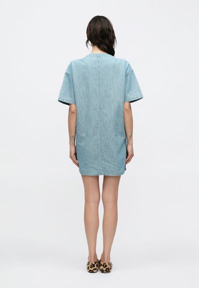 Γυναίκα που στέκεται πλάτη, φοράει κοντομάνικο, γαλάζιο φόρεμα από τζιν και παπούτσια slip-on με λεοπάρ σχέδιο πάνω σε λευκό φόντο.