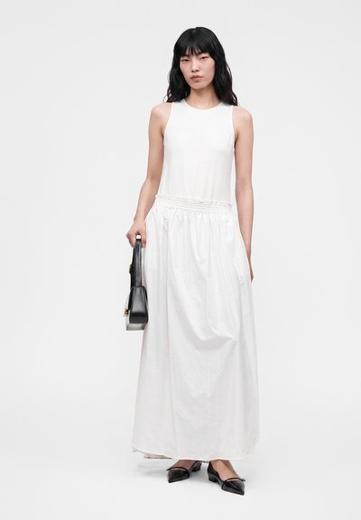 Femme portant une robe longue blanche sans manches, tenant un sac à main noir, debout devant un fond clair uni.