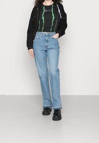 Black bomber jacket with white accents, green tie-dye fitted top, light blue high-waisted jeans, and chunky black shoes.