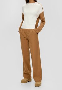 Femme portant un pull à manches longues blanc et marron, un pantalon large marron et des chaussures à plateforme tan avec les mains dans les poches.
