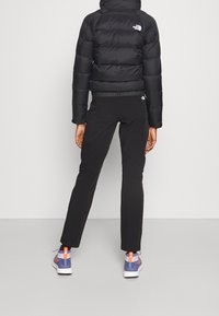 Veste matelassée noire avec un quilting horizontal et un col haut, associée à un pantalon noir et des chaussures de sport multicolores.