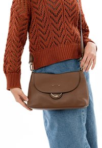 Brun læder skuldertaske med en afrundet klap, sølvbeslag og tekstureret overflade, holdt af en person iført en orange strikket sweater.