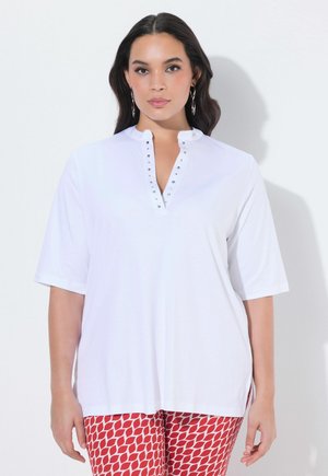 Frau mit langen dunklen Haaren trägt ein weißes Kurzarmshirt mit Nieten am V-Ausschnitt und rote gemusterte Hosen, steht vor hellem Hintergrund.